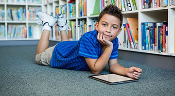 A boy on his stomach with a tablet on carpeted library floor